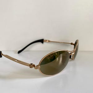 May include: Gold-tone Ray-Ban sunglasses with oval lenses. The sunglasses have a gold frame, brown lenses, and black earpieces. The Ray-Ban logo is visible on the lens.