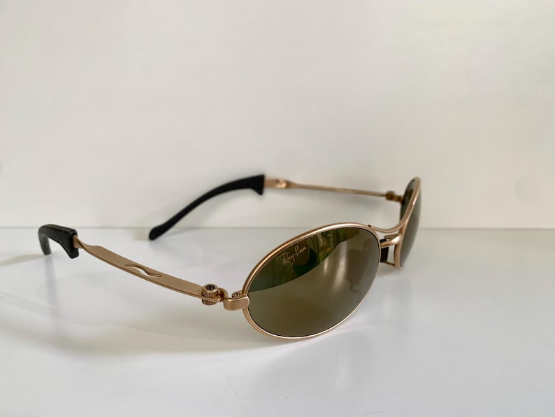 May include: Gold-tone Ray-Ban sunglasses with oval lenses. The sunglasses have a gold frame, brown lenses, and black earpieces. The Ray-Ban logo is visible on the lens.