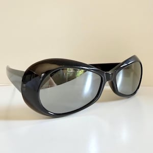 May include: Black sunglasses with a thick, rounded frame and reflective lenses. The sunglasses have a sleek, modern design, with a curved shape and a glossy finish. The lenses are a mirrored silver colour.