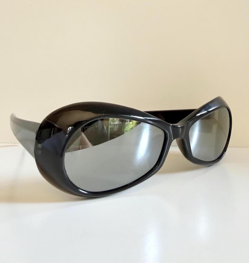 May include: Black sunglasses with a thick, rounded frame and reflective lenses. The sunglasses have a sleek, modern design, with a curved shape and a glossy finish. The lenses are a mirrored silver colour.