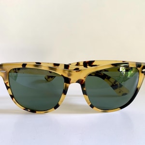 May include: Tortoiseshell sunglasses with dark green lenses. The frames have a classic, retro design with a speckled pattern in shades of tan and brown. The sunglasses are set against a white background.