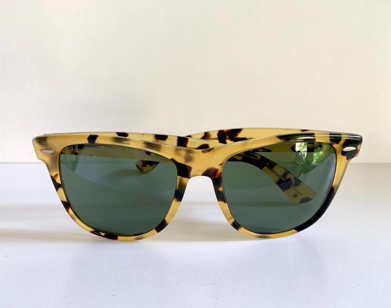 May include: Tortoiseshell sunglasses with dark green lenses. The frames have a classic, retro design with a speckled pattern in shades of tan and brown. The sunglasses are set against a white background.