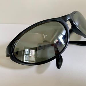May include: Black sunglasses with a wrap-around frame design and dark tinted lenses. The left lens features the white Ray-Ban logo printed in the upper corner. The frames are constructed from a smooth, matte black material with curved temples.