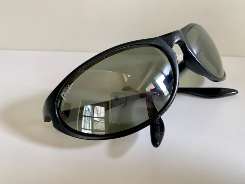 May include: Black sunglasses with a wrap-around frame design and dark tinted lenses. The left lens features the white Ray-Ban logo printed in the upper corner. The frames are constructed from a smooth, matte black material with curved temples.