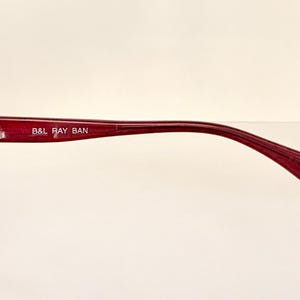 May include: Close-up of a pair of burgundy sunglasses. The frame is a deep red color with a sleek, modern design. The text "B&L RAY BAN" is printed on the temple arm. The lenses are a reflective silver color.