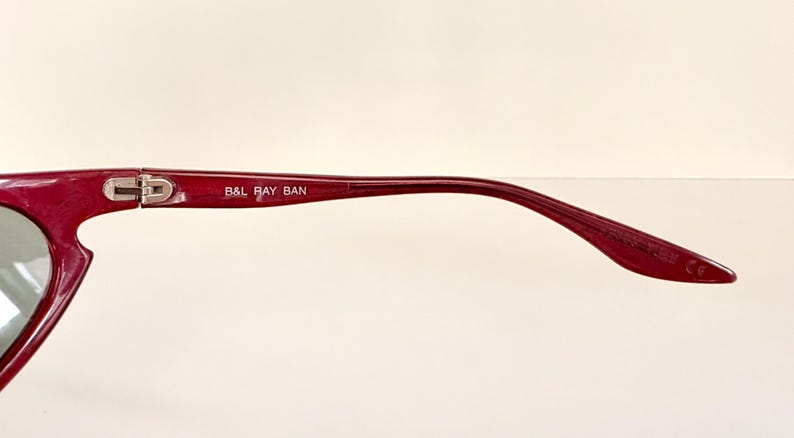May include: Close-up of a pair of burgundy sunglasses. The frame is a deep red color with a sleek, modern design. The text "B&L RAY BAN" is printed on the temple arm. The lenses are a reflective silver color.