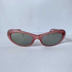 Peut inclure: Lunettes de soleil &agrave; monture rose avec verres gris. Les lunettes de soleil ont une forme &oelig;il de chat et le logo Ray-Ban est visible sur la lentille gauche. La monture est de couleur rose translucide.