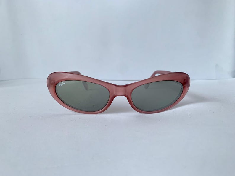Peut inclure: Lunettes de soleil &agrave; monture rose avec verres gris. Les lunettes de soleil ont une forme &oelig;il de chat et le logo Ray-Ban est visible sur la lentille gauche. La monture est de couleur rose translucide.