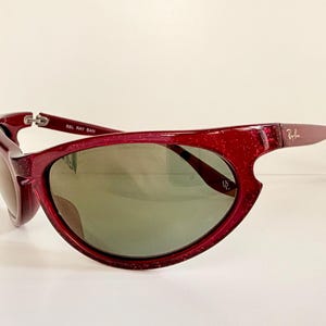 May include: A pair of red-framed sunglasses with a glittery finish. The lenses are a muted green color. The Ray-Ban logo is visible on the right arm. The sunglasses are set against a white background.