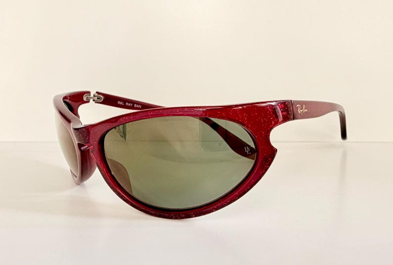 May include: A pair of red-framed sunglasses with a glittery finish. The lenses are a muted green color. The Ray-Ban logo is visible on the right arm. The sunglasses are set against a white background.