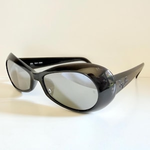 May include: Black sunglasses with a rounded, oversized frame and silver lenses. The sunglasses have a glossy finish and the Ray-Ban logo is visible on the temple. The sunglasses are set against a white background.