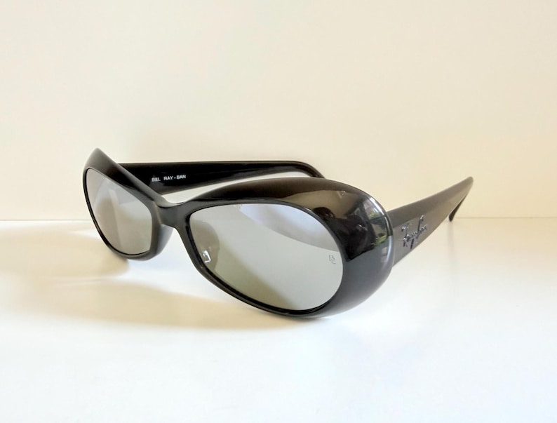 May include: Black sunglasses with a rounded, oversized frame and silver lenses. The sunglasses have a glossy finish and the Ray-Ban logo is visible on the temple. The sunglasses are set against a white background.