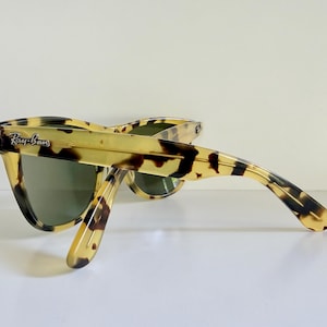 May include: Tortoiseshell-patterned sunglasses with green lenses. The Ray-Ban logo is visible on the left temple. The sunglasses are angled, showcasing the frame's design and the lenses' tint. The frame has a classic, retro style.