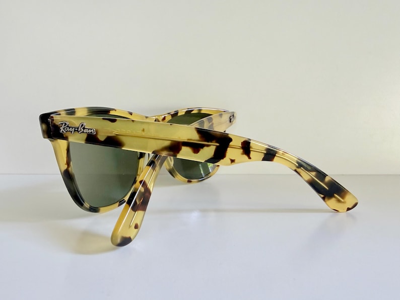 May include: Tortoiseshell-patterned sunglasses with green lenses. The Ray-Ban logo is visible on the left temple. The sunglasses are angled, showcasing the frame's design and the lenses' tint. The frame has a classic, retro style.