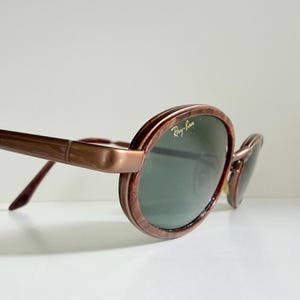 May include: Brown oval sunglasses with green lenses. The Ray-Ban logo is visible on the lens. The frames have a marbled effect. The arms are a solid brown colour. The sunglasses are angled slightly.