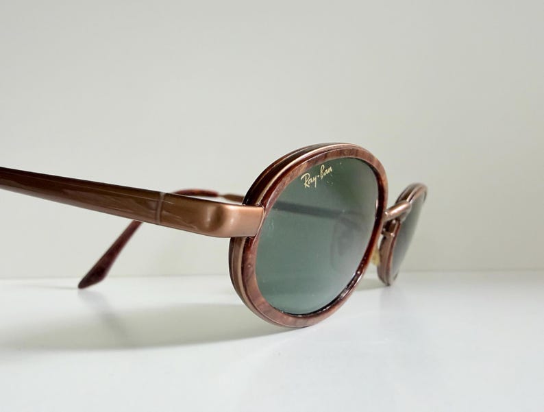 May include: Brown oval sunglasses with green lenses. The Ray-Ban logo is visible on the lens. The frames have a marbled effect. The arms are a solid brown colour. The sunglasses are angled slightly.