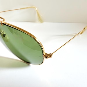 May include: Gold-framed sunglasses with green lenses. The sunglasses have a classic aviator design with a double bridge and a gold metal frame. The lenses are a light green color, and the arms are thin and gold. The sunglasses are set against a white background.