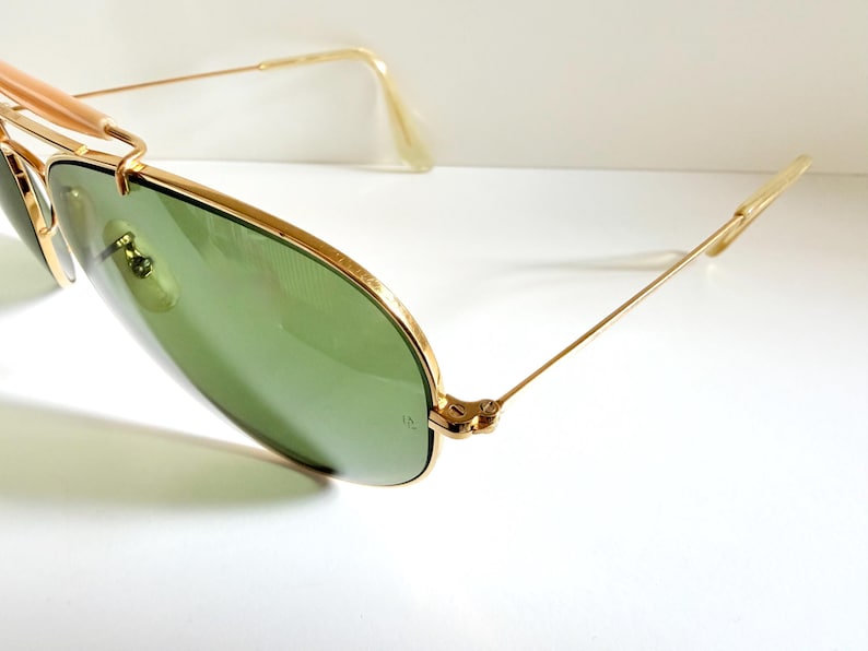 May include: Gold-framed sunglasses with green lenses. The sunglasses have a classic aviator design with a double bridge and a gold metal frame. The lenses are a light green color, and the arms are thin and gold. The sunglasses are set against a white background.