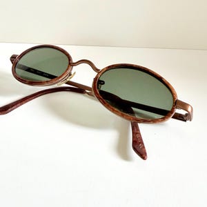 May include: Oval sunglasses with a brown frame and green lenses. The sunglasses have a classic design with a thin metal frame and a curved bridge. The arms are a darker brown colour. The lenses are a dark green colour.