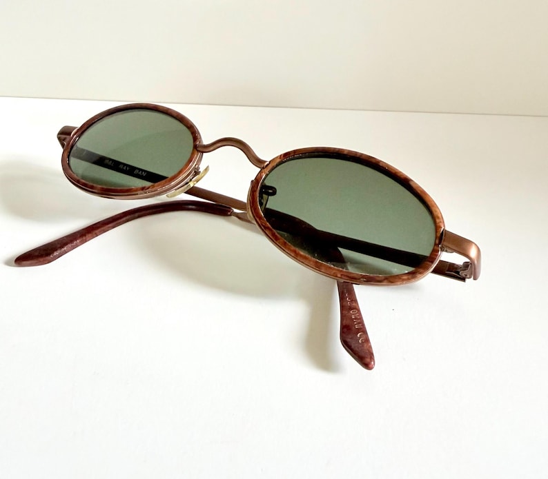 May include: Oval sunglasses with a brown frame and green lenses. The sunglasses have a classic design with a thin metal frame and a curved bridge. The arms are a darker brown colour. The lenses are a dark green colour.