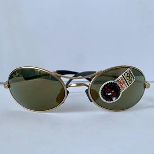 May include: Gold-framed sunglasses with oval lenses and a black bridge. The lenses are a dark green color. A sticker on the right lens reads "SPORT" and "88". The sunglasses have a classic, sporty design.