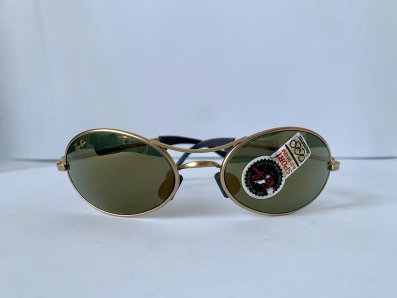 May include: Gold-framed sunglasses with oval lenses and a black bridge. The lenses are a dark green color. A sticker on the right lens reads "SPORT" and "88". The sunglasses have a classic, sporty design.
