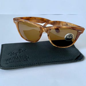 Puede incluir: Gafas de sol Ray-Ban Wayfarer de carey con lentes marrones y una funda de cuero negro. La funda tiene el logotipo de Ray-Ban y las palabras "Wayfarer by Bausch & Lomb" impresas en ella.