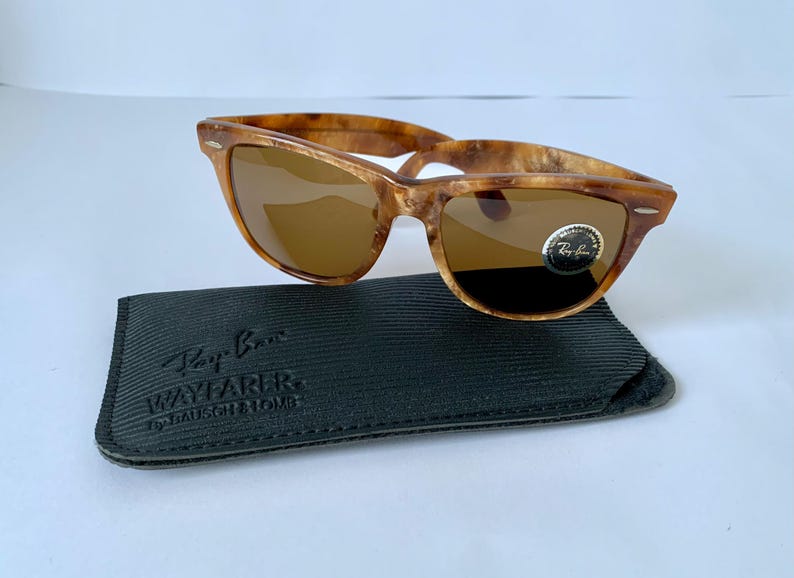 Puede incluir: Gafas de sol Ray-Ban Wayfarer de carey con lentes marrones y una funda de cuero negro. La funda tiene el logotipo de Ray-Ban y las palabras "Wayfarer by Bausch & Lomb" impresas en ella.