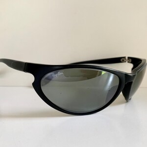 May include: Black wrap-around sunglasses featuring a matte black frame and dark grey tinted lenses. The left lens displays the white Ray-Ban logo in the upper corner. These sunglasses have a curved, aerodynamic shape with thin temples.