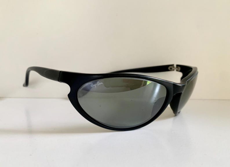 May include: Black wrap-around sunglasses featuring a matte black frame and dark grey tinted lenses. The left lens displays the white Ray-Ban logo in the upper corner. These sunglasses have a curved, aerodynamic shape with thin temples.