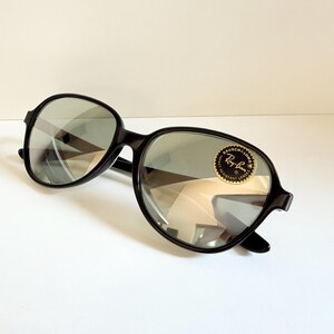 May include: Black Ray-Ban sunglasses with oval lenses. The sunglasses have a black frame and a gold and black sticker on the lens. The lenses reflect the surrounding environment.
