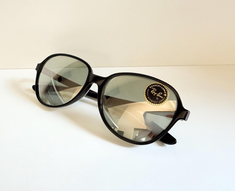 May include: Black Ray-Ban sunglasses with oval lenses. The sunglasses have a black frame and a gold and black sticker on the lens. The lenses reflect the surrounding environment.