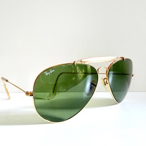 May include: Gold-framed aviator sunglasses with green lenses. The Ray-Ban logo is visible on the lens. The sunglasses have a double bridge and a white brow bar. The temples are thin and gold-colored.