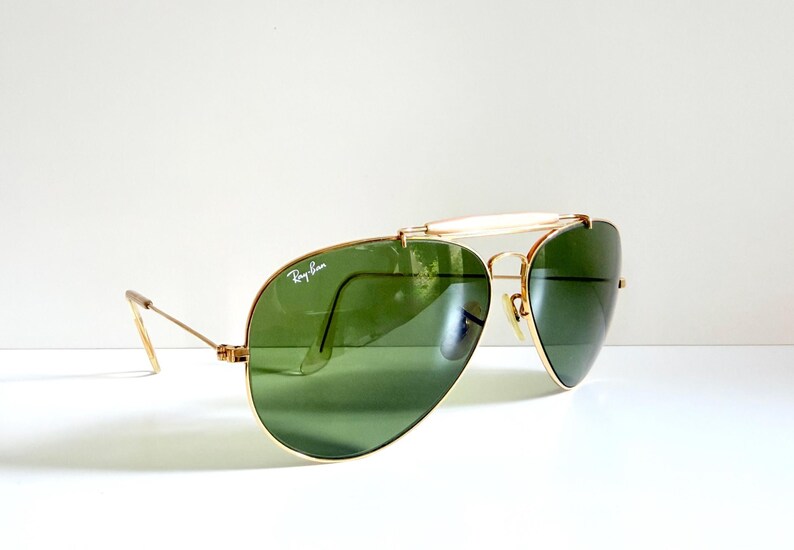 May include: Gold-framed aviator sunglasses with green lenses. The Ray-Ban logo is visible on the lens. The sunglasses have a double bridge and a white brow bar. The temples are thin and gold-colored.
