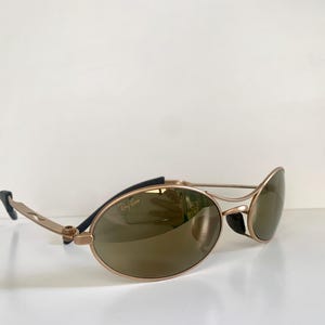 May include: Gold-tone sunglasses with oval lenses and a sleek, modern design. The sunglasses feature a gold frame with black accents on the temples and a subtle Ray-Ban logo. The lenses are a warm brown color.