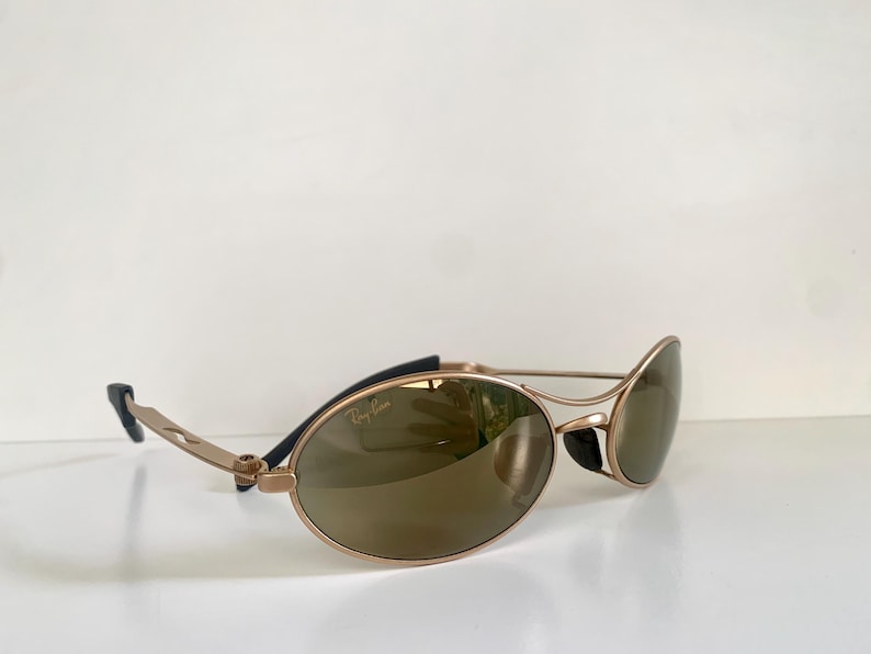 May include: Gold-tone sunglasses with oval lenses and a sleek, modern design. The sunglasses feature a gold frame with black accents on the temples and a subtle Ray-Ban logo. The lenses are a warm brown color.