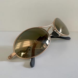 May include: Gold-framed sunglasses with oval lenses. The lenses are a dark green color, and the frames have a classic aviator design. The sunglasses are resting on a white surface.