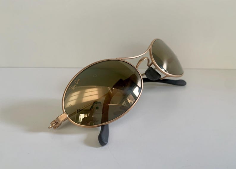 May include: Gold-framed sunglasses with oval lenses. The lenses are a dark green color, and the frames have a classic aviator design. The sunglasses are resting on a white surface.