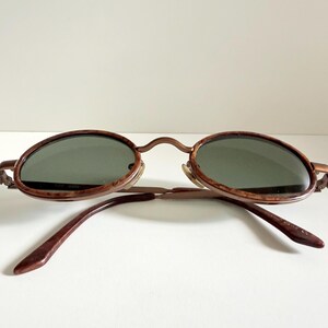 May include: Vintage oval sunglasses with a brown tortoiseshell frame and dark green lenses. The arms are a matching brown colour. The sunglasses are set against a white background.