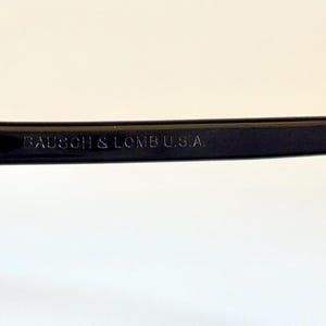 May include: Close-up of a black-framed pair of sunglasses with green lenses. The text "BAUSCH & LOMB U.S.A." is printed on the temple arm. The glasses have a silver hinge and a classic design.