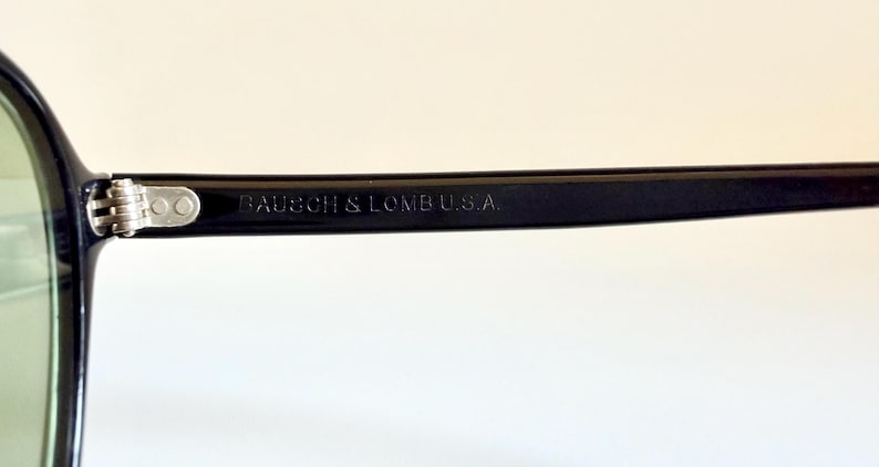 May include: Close-up of a black-framed pair of sunglasses with green lenses. The text "BAUSCH & LOMB U.S.A." is printed on the temple arm. The glasses have a silver hinge and a classic design.