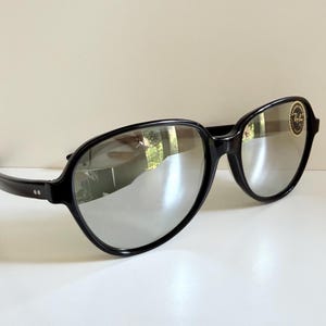 May include: Black sunglasses with silver mirrored lenses. The frames are thick and rounded, with a small logo on the right lens. The sunglasses are resting on a white surface.