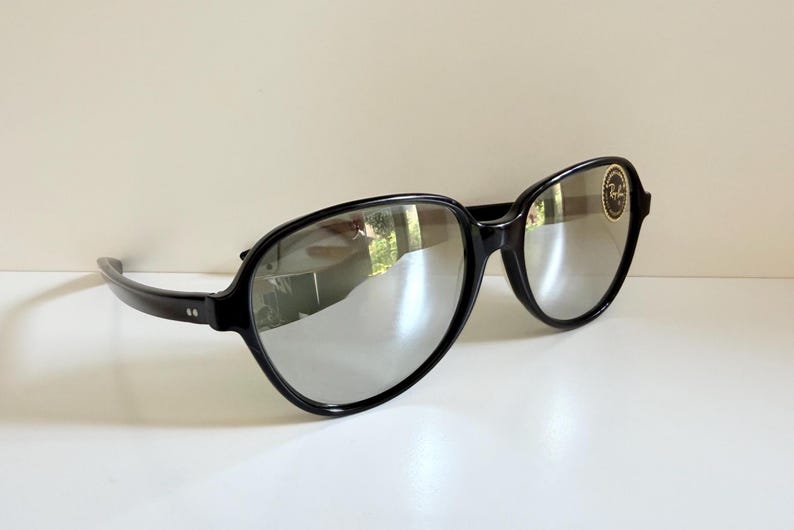 May include: Black sunglasses with silver mirrored lenses. The frames are thick and rounded, with a small logo on the right lens. The sunglasses are resting on a white surface.