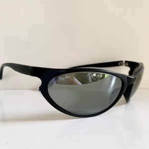 May include: Black sunglasses with a wrap-around frame design and reflective silver-toned lenses. The left lens features the white Ray-Ban logo printed on the upper corner. The frames are matte black with curved temples and a sturdy bridge construction.