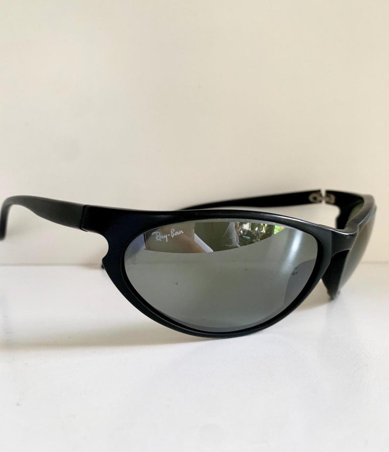 May include: Black sunglasses with a wrap-around frame design and reflective silver-toned lenses. The left lens features the white Ray-Ban logo printed on the upper corner. The frames are matte black with curved temples and a sturdy bridge construction.