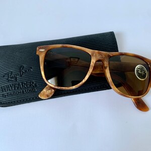 Puede incluir: Gafas de sol Ray-Ban Wayfarer de carey con lentes marrones en una funda negra. La funda tiene el logotipo de Ray-Ban Wayfarer y el texto "By Bausch & Lomb".