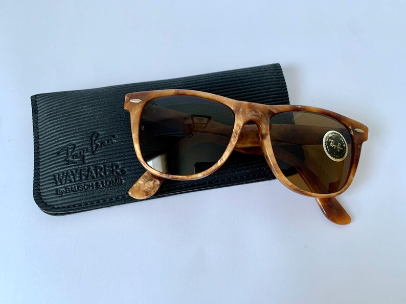 Puede incluir: Gafas de sol Ray-Ban Wayfarer de carey con lentes marrones en una funda negra. La funda tiene el logotipo de Ray-Ban Wayfarer y el texto "By Bausch & Lomb".