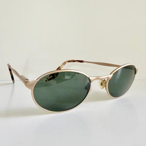 Puede incluir: Gafas de sol ovaladas doradas con lentes verdes. Las gafas tienen una montura met&aacute;lica fina y un logotipo de Ray-Ban en la lente izquierda. Las patillas tienen un patr&oacute;n de carey. Sobre una superficie blanca.