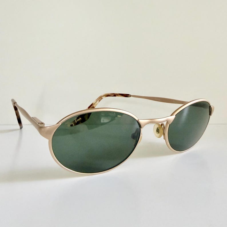 Puede incluir: Gafas de sol ovaladas doradas con lentes verdes. Las gafas tienen una montura met&aacute;lica fina y un logotipo de Ray-Ban en la lente izquierda. Las patillas tienen un patr&oacute;n de carey. Sobre una superficie blanca.
