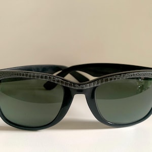 May include: Black sunglasses with a textured, dark gray upper frame and dark green lenses. The sunglasses have a classic, retro design, suitable for both men and women. The frames are made of a glossy black material.